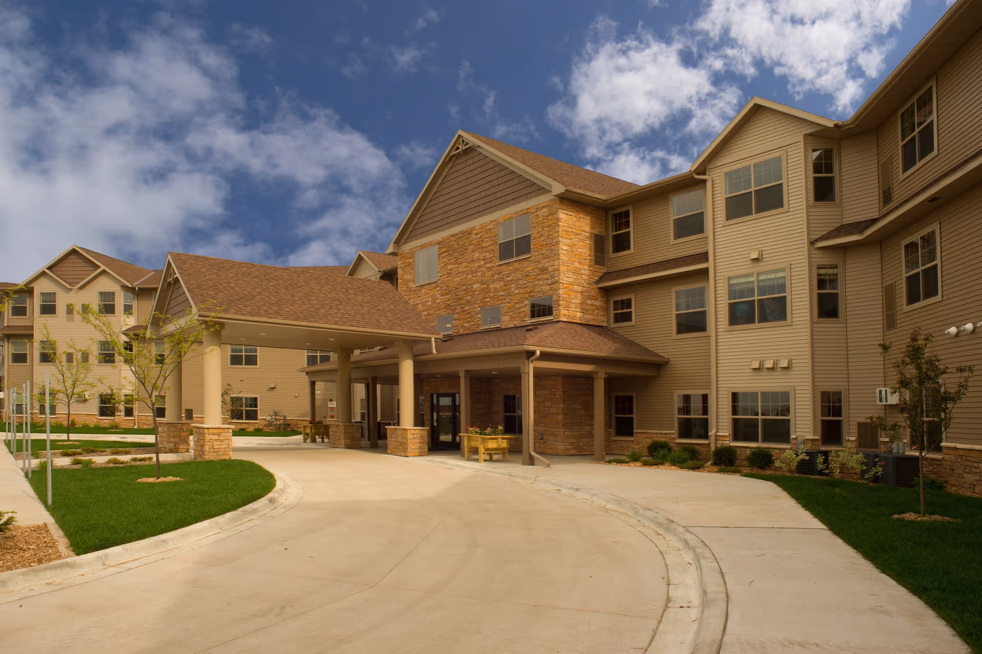 Exterior view of a senior living facility building with beige siding and stone accents under a partly cloudy sky. The building has multiple windows and a covered entrance with pillars. There is a curved driveway and landscaped green areas with young trees and shrubs.