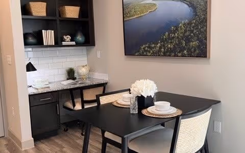 Small dining area with a black table set for two, cane-back chairs, a built-in desk nook with shelves and decorative items, and a large landscape wall art.