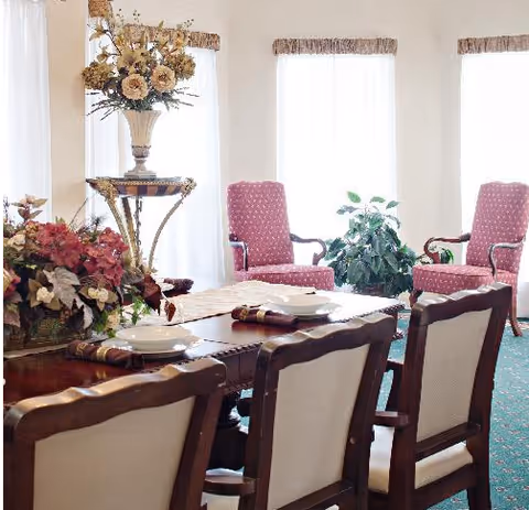 A formal dining room with a wooden table set with plates and napkins, upholstered chairs, floral centerpieces, and armchairs by tall sunlit windows.