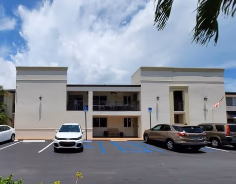 Exterior view of a two-story senior living facility building with a parking lot in front. Several cars are parked in marked spaces, including handicapped spots. The building is light-colored with balconies on the upper floor and an American flag on the right side. Palm tree leaves are visible at the top right corner under a partly cloudy sky.