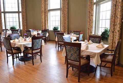 Bright dining room with several small tables set with white tablecloths and wooden chairs by large windows and patterned curtains.
