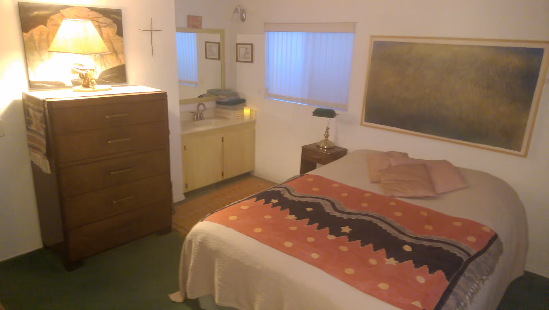 A small bedroom featuring a bed with a patterned throw, a wooden dresser with a lamp, a sink vanity, and framed artwork on the walls.