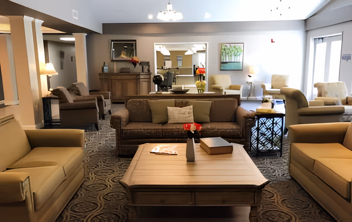A spacious and well-lit living room area with multiple sofas and armchairs arranged around a central wooden coffee table. The room features patterned carpeting, table lamps, decorative pillows, a side table with a flower vase, and artwork on the walls. Large windows allow natural light to brighten the space.