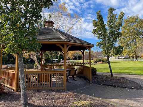 A wooden gazebo with benches inside situated in a landscaped outdoor area with trees, grass, and a paved walkway under a partly cloudy blue sky.