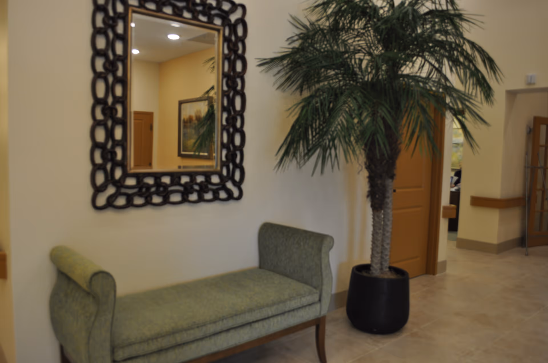 Indoor seating area with a green upholstered bench, a large decorative mirror with a chain-link frame on the wall, and a tall potted palm tree next to a doorway in a hallway with beige walls and tiled floor.
