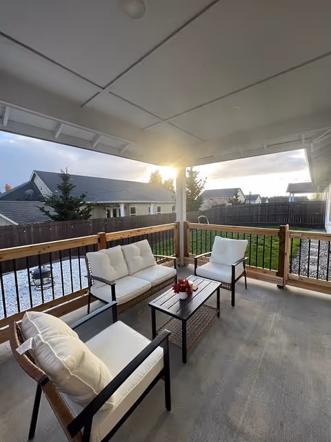 Covered outdoor patio area with cushioned seating including a sofa and two chairs around a small coffee table with a red flower centerpiece. The patio overlooks a fenced backyard with grass and neighboring houses visible in the background during sunset.