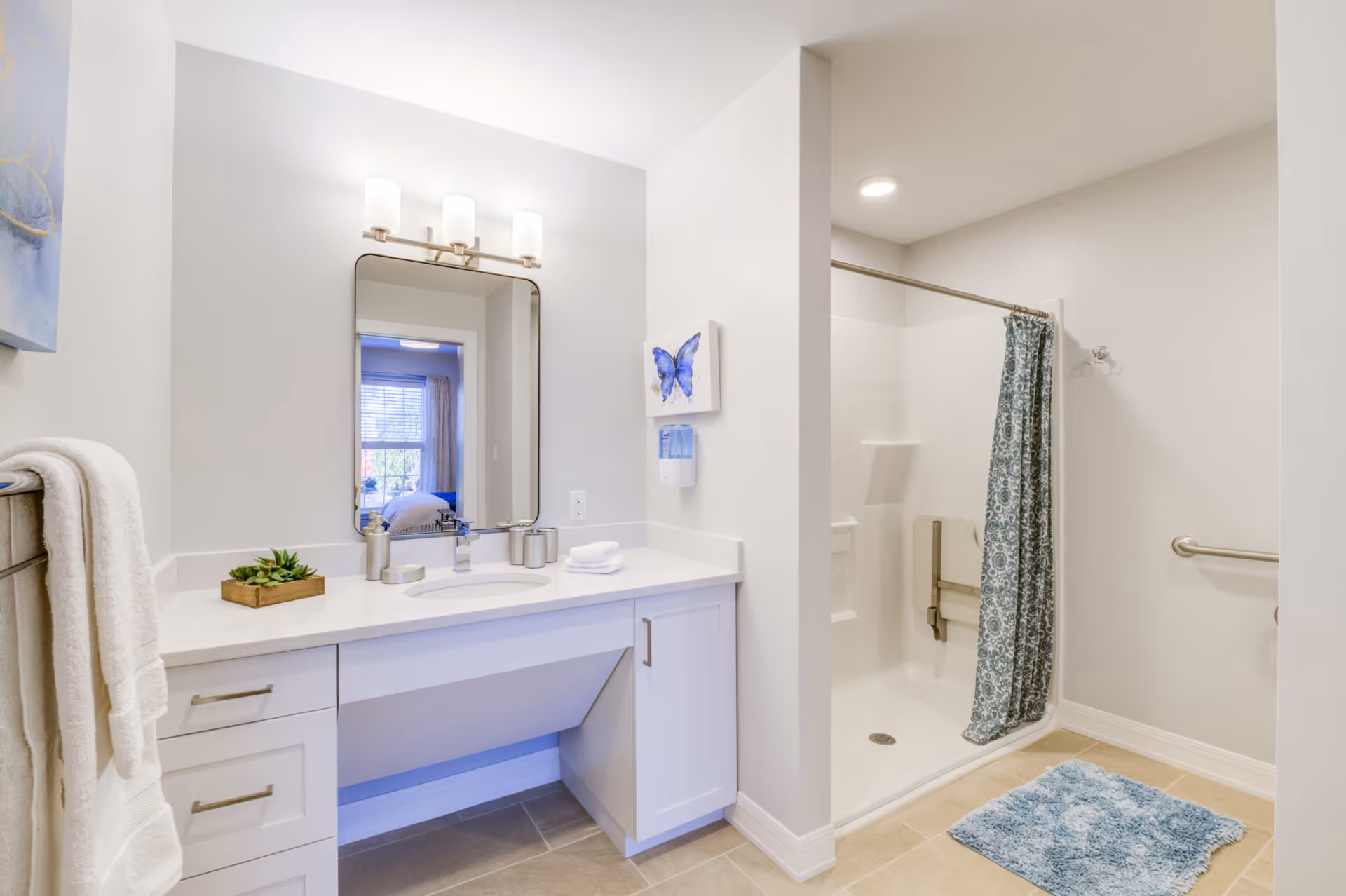A bright and clean bathroom featuring a white vanity with a sink, a large mirror with three light fixtures above it, a towel rack with a white towel, a small plant on the counter, and a walk-in shower with a patterned blue shower curtain and grab bars. A blue bath mat is placed on the floor in front of the shower.