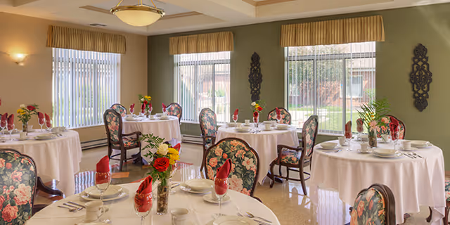 A bright dining room with round tables covered in white tablecloths, each set with plates, cups, and red folded napkins. Floral upholstered chairs surround the tables. Large windows with vertical blinds and valances let in natural light, and decorative wall hangings and flower arrangements add to the cozy atmosphere.