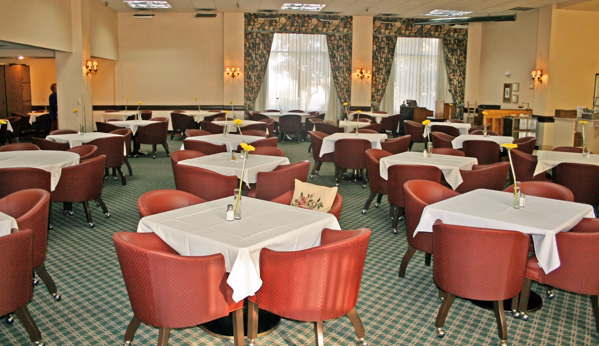A dining room with multiple square tables covered with white tablecloths, each surrounded by red upholstered chairs. Each table has a single yellow flower in a vase and salt and pepper shakers. The room has large windows with floral curtains and wall sconces providing warm lighting. The carpet is green with a patterned design.
