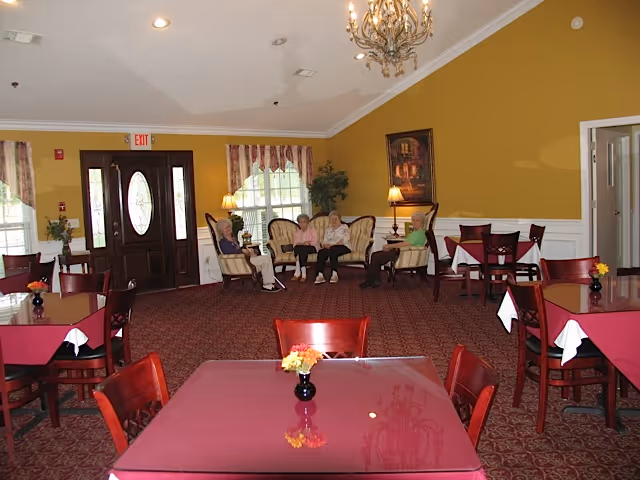 A spacious dining room with several tables covered in red tablecloths and small flower vases. Four elderly people are seated on upholstered chairs and a sofa near the back wall, which is painted yellow. There are two windows with curtains, a chandelier hanging from the ceiling, and a dark wooden door with an oval glass panel.
