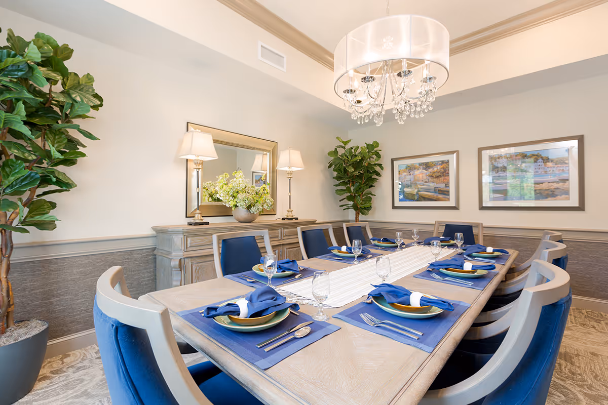 A well-lit dining room with a long wooden table set for eight people. Each place setting includes a blue placemat, a plate, a blue cloth napkin with a white napkin ring, a fork, a knife, and a water glass. The room features two large potted plants, a sideboard with two lamps and a flower arrangement, a large mirror, two framed landscape paintings on the wall, and a chandelier with crystal accents hanging above the table.