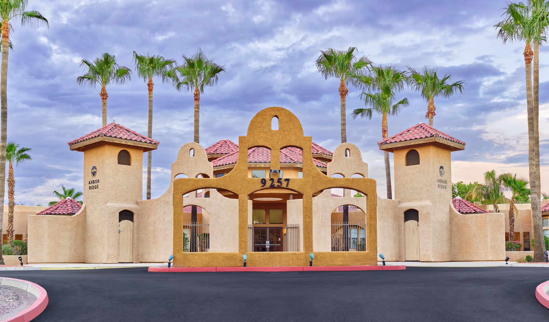 Front exterior view of Arbor Ridge facility with a distinctive southwestern architectural style, featuring tan stucco walls, red tile roofs, palm trees, and a large decorative archway with the number 9257 displayed above the entrance.