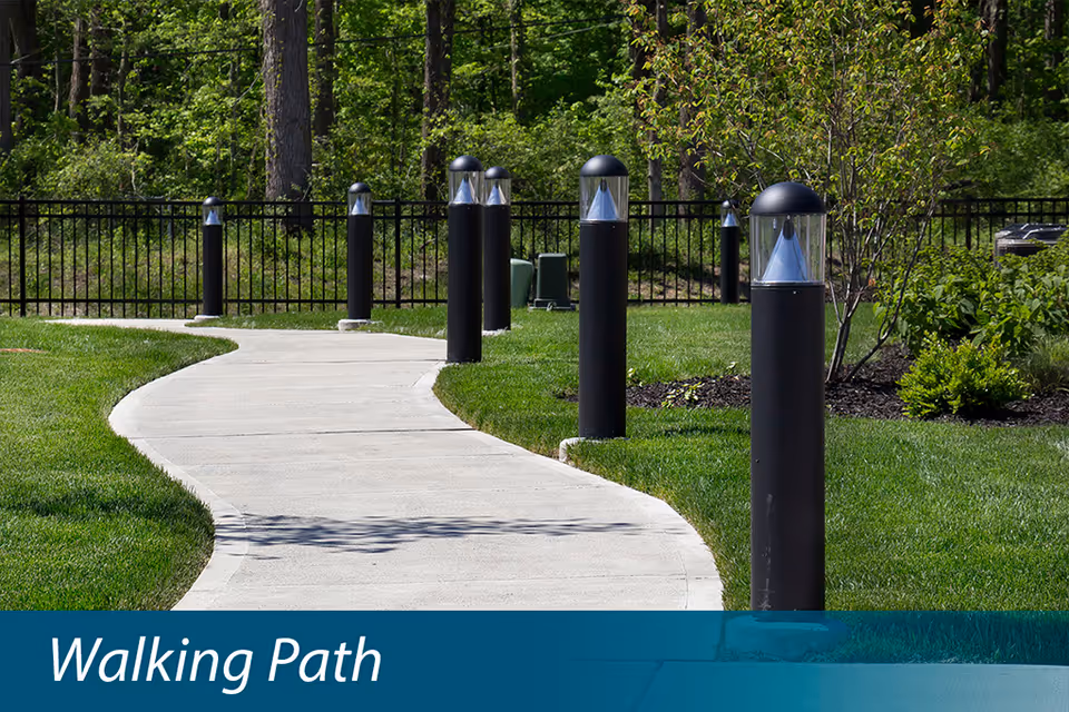 Curved concrete walking path lined with black bollard lights through a grassy, tree-lined landscaped area.