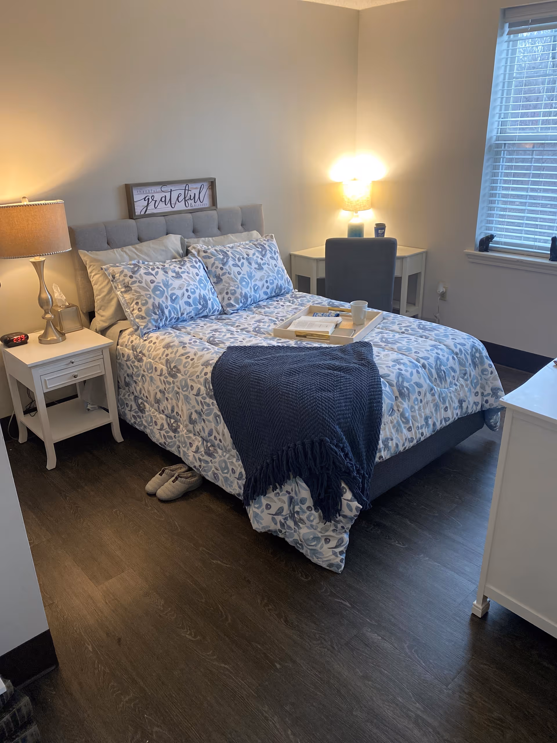 A cozy bedroom with a neatly made bed featuring blue and white floral bedding and a dark blue throw blanket. There is a white nightstand with a lamp and an alarm clock on the left side of the bed. A pair of slippers is placed on the floor beside the bed. On the right side, there is a small white desk with a chair and a lit lamp. A window with blinds is on the right wall, allowing natural light into the room. A decorative sign above the bed reads 'grateful'.