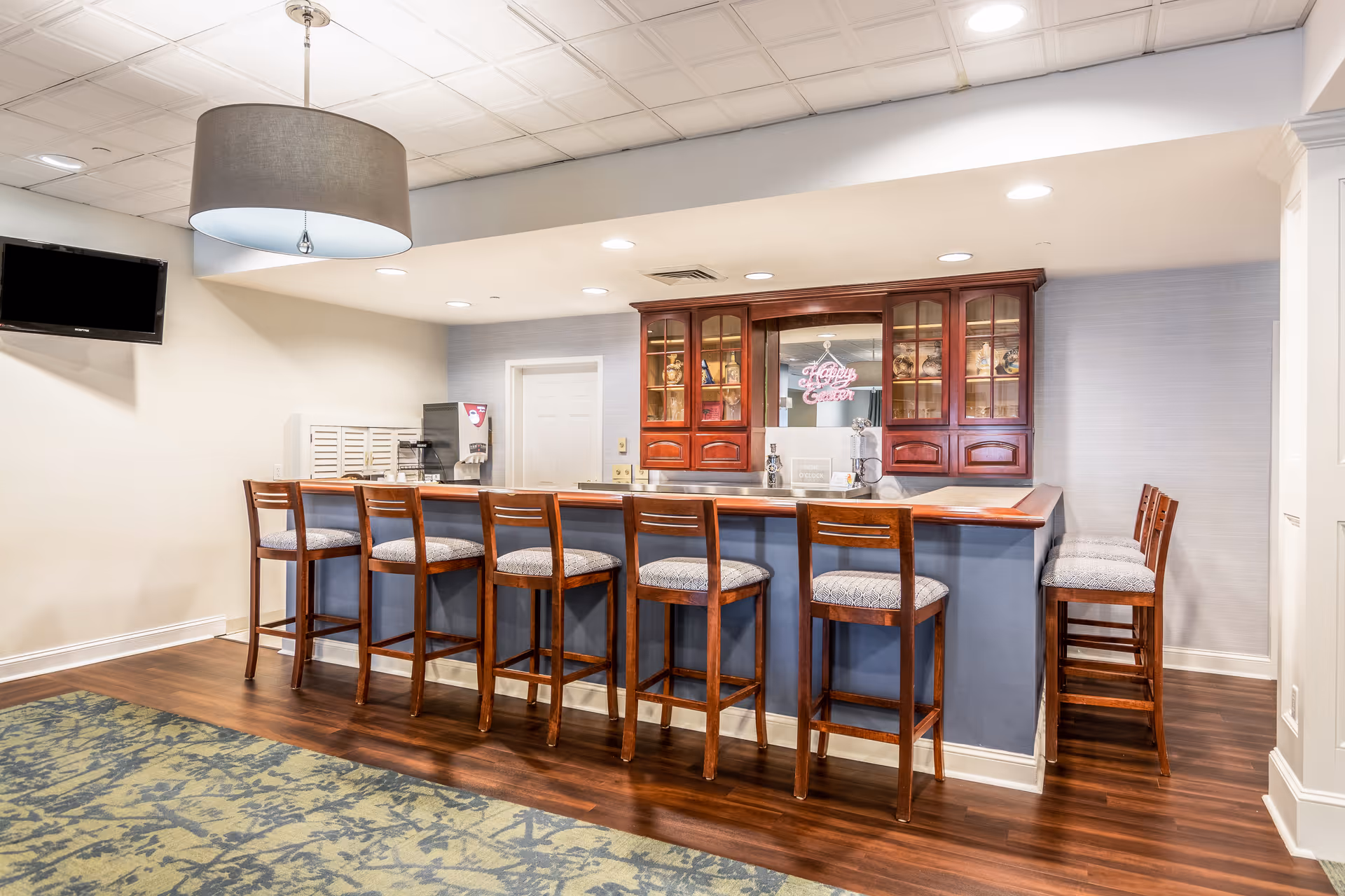 Interior view of a seating area with a long counter and eight wooden bar stools with cushioned seats. Behind the counter is a cabinet with glass doors displaying decorative items and a neon sign that reads 'Happy Easter'. The floor is wooden with a patterned carpet in front of the counter, and a large ceiling light fixture hangs above. A wall-mounted TV is visible on the left side.