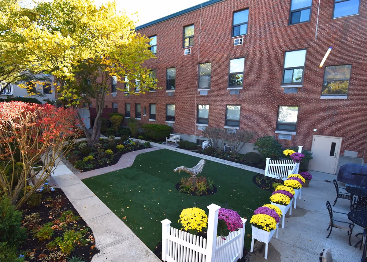 Outdoor garden area at New Horizons At Choate featuring a green lawn with a small tree stump centerpiece, surrounded by flower beds and colorful potted flowers in white planters. There are benches and black metal tables with chairs on a concrete patio next to a red brick building with multiple windows.