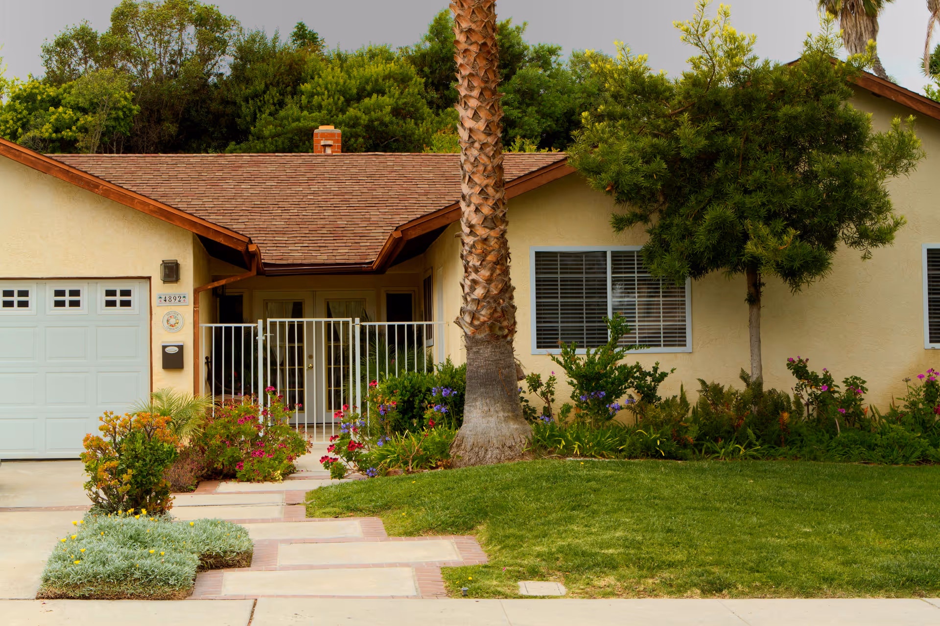 Front exterior view of a single-story residential building with a brown shingle roof, beige walls, a white garage door, a gated entrance, and a well-maintained garden with green grass, colorful flowers, and trees.