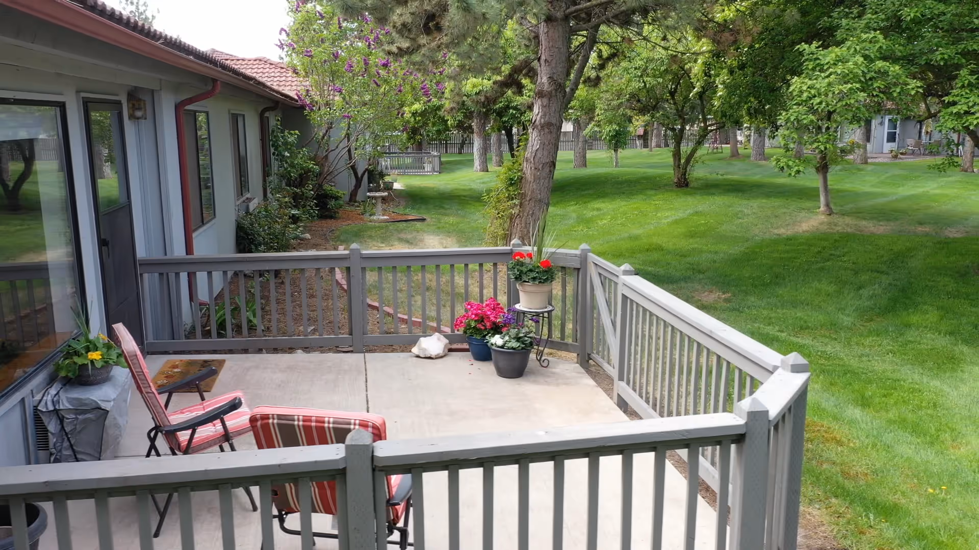 A small fenced patio area with two chairs and several potted plants, adjacent to a building with large windows. Beyond the patio is a well-maintained grassy area with trees and shrubs.