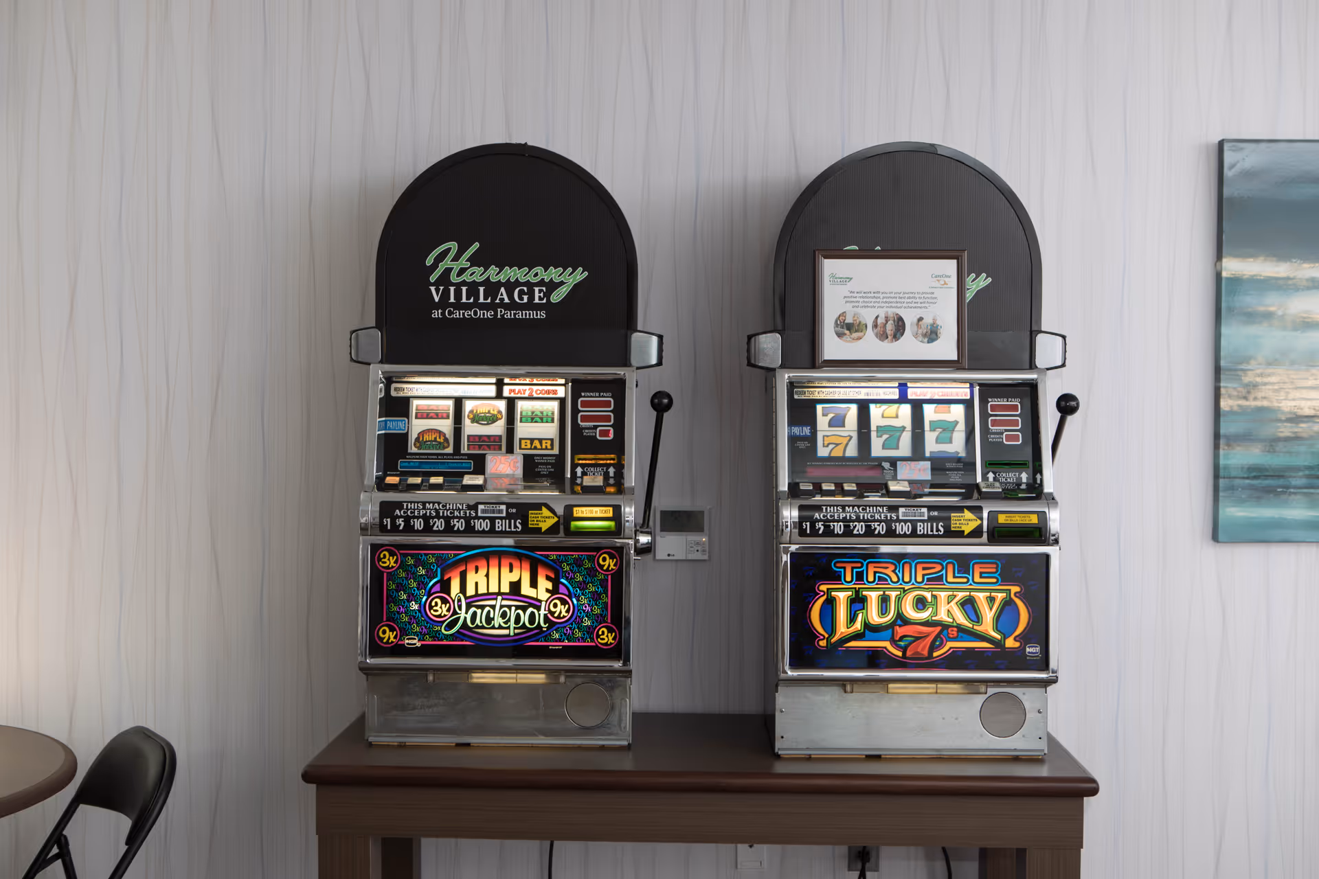 Two vintage slot machines labeled Triple Jackpot and Triple Lucky placed side by side on a wooden table against a light-colored wall with subtle vertical patterns. A portion of a chair and a round table are visible on the left, and a framed abstract painting is partially visible on the right.