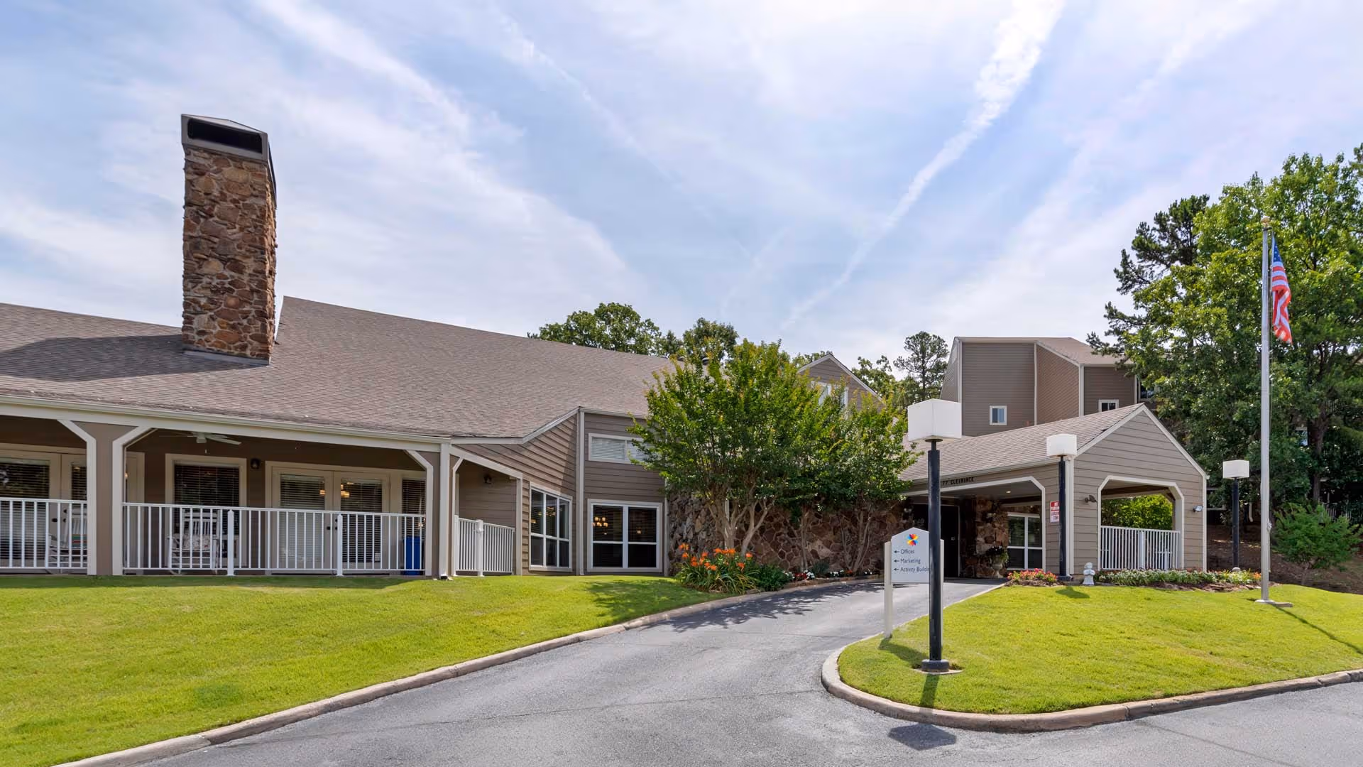 Front entrance of a senior living building with a covered porte-cochère, wraparound porch, stone chimney, and flagpole on a grassy driveway.