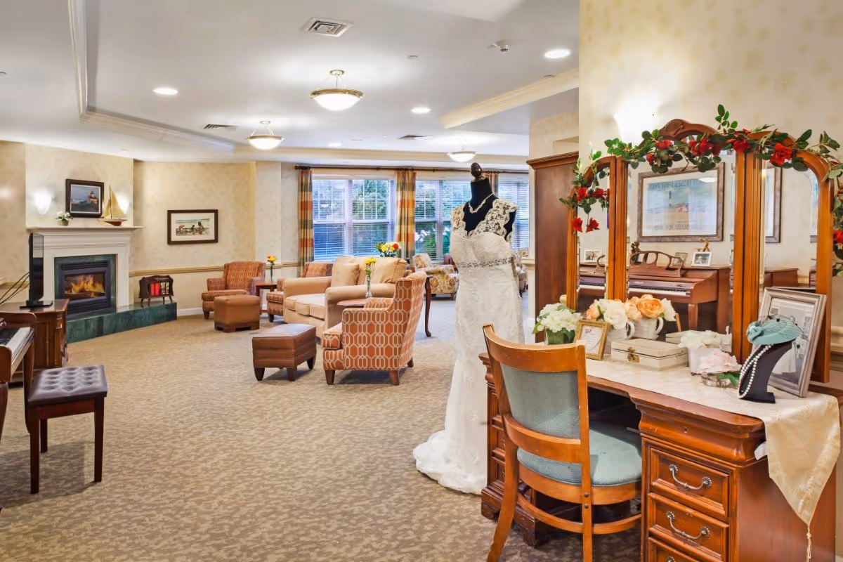 Spacious senior living lounge with sofas and armchairs around a fireplace and a vanity displaying a wedding dress on a mannequin.