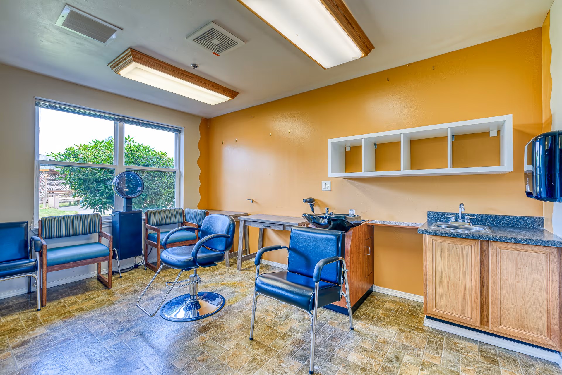 Interior view of a small salon or barber area in an assisted living facility with two black salon chairs, a hair washing station, a countertop with a sink, and several waiting chairs near a large window showing greenery outside. The walls are painted yellow and the floor has a patterned tile.