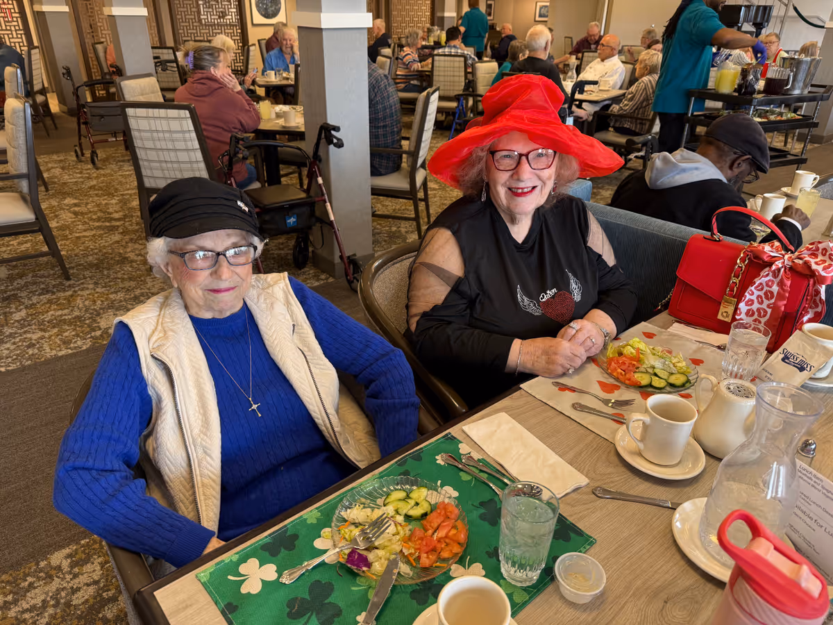Two elderly women sitting at a dining table in a senior living facility. One woman is wearing a black hat, glasses, a blue sweater, and a white vest, while the other woman is wearing a bright red hat, glasses, and a black top with a heart design. Both have plates with salad in front of them. Other residents are visible in the background seated at tables, and a staff member is serving drinks.