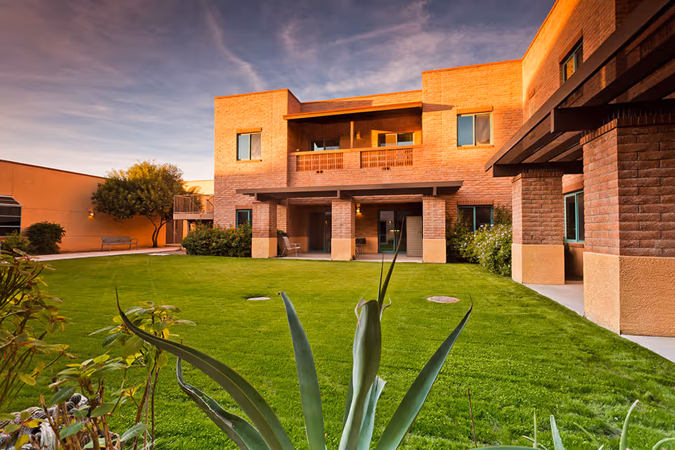 A well-maintained outdoor courtyard area of a senior living facility with green grass, plants, and a two-story brick building in the background under a partly cloudy sky during sunset.