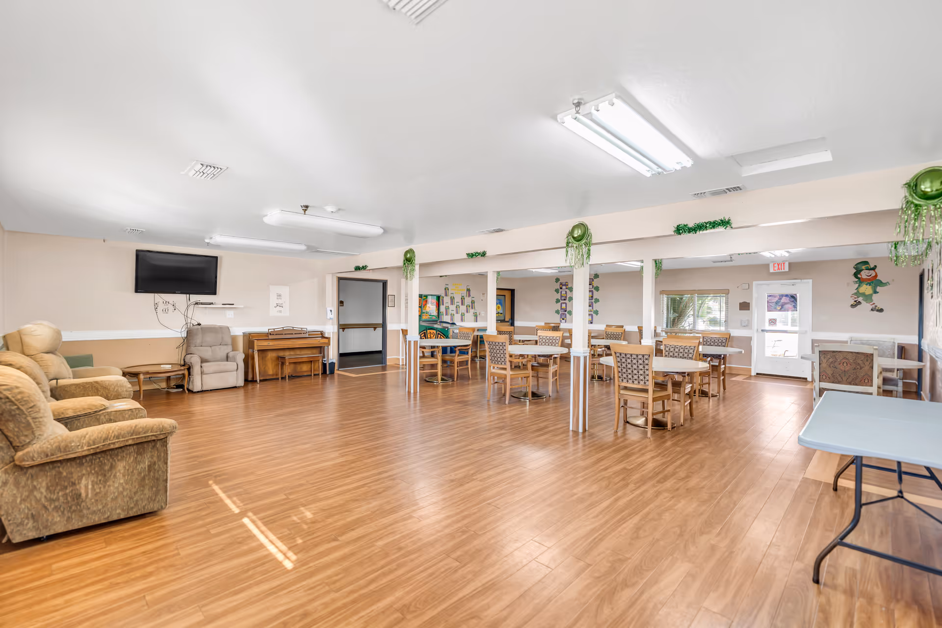 A spacious common room with wooden flooring, several round tables with chairs, a few armchairs, a wall-mounted TV, and a piano. The room is decorated with green hanging decorations and has fluorescent ceiling lights. There is a door and windows on the far wall.