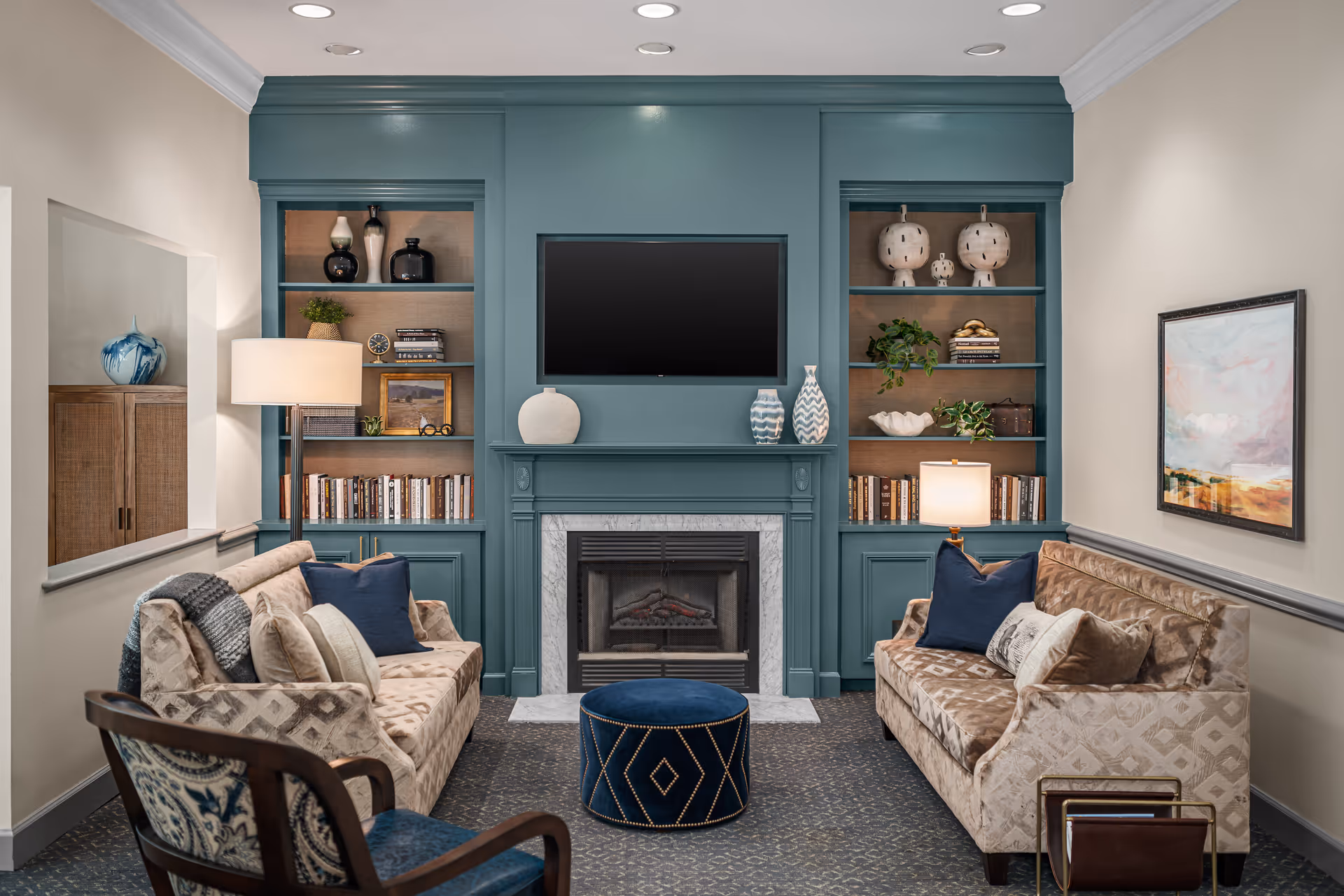 A cozy living room with two patterned sofas facing each other, a blue ottoman in the center, and a fireplace with a mounted flat-screen TV above it. Built-in shelves on either side of the fireplace hold decorative vases, books, plants, and lamps. A framed painting hangs on the right wall, and a wooden chair with blue upholstery is partially visible in the foreground.