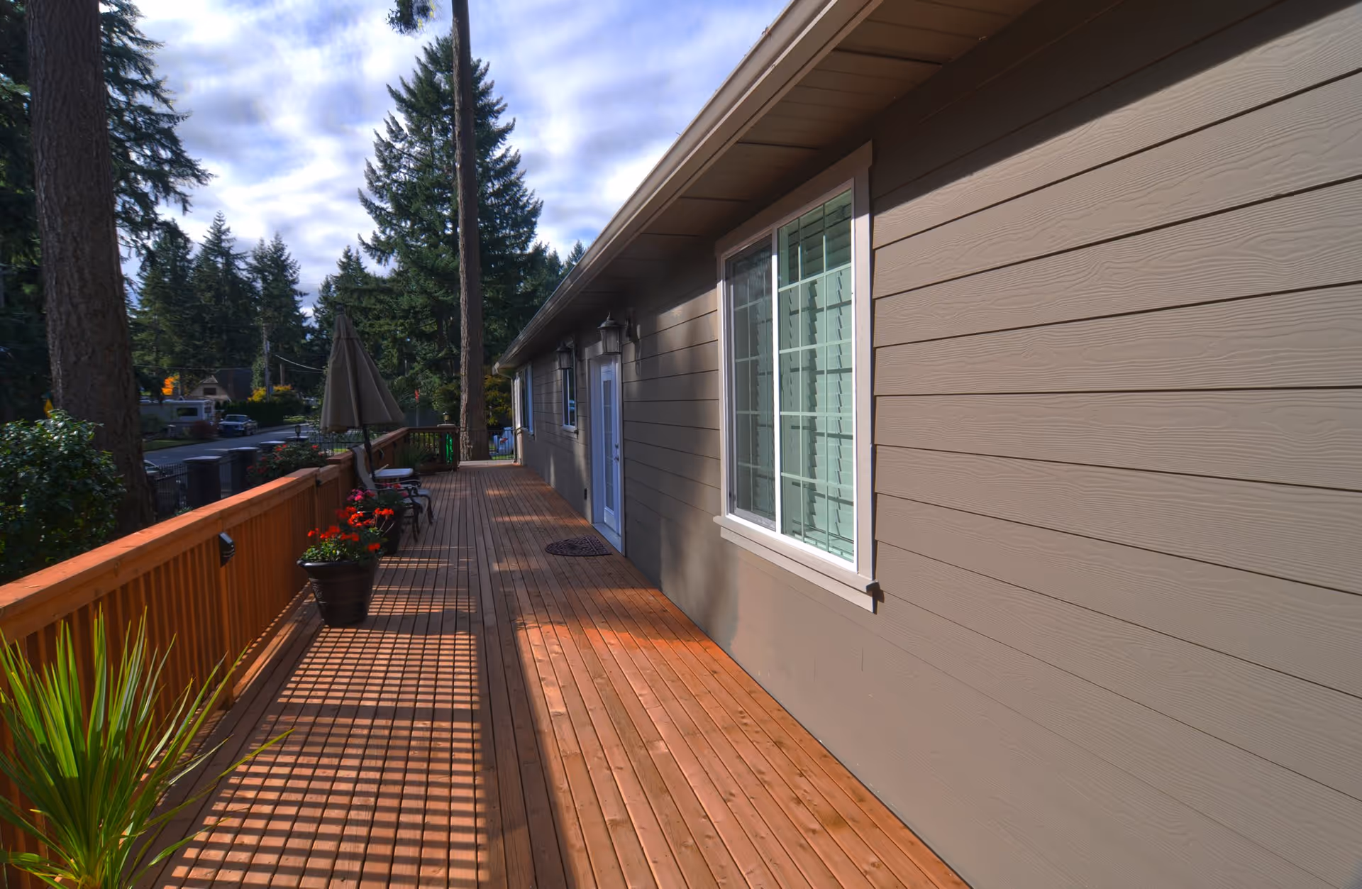 A wooden deck attached to the side of a building with beige siding and white-framed windows. The deck has a wooden railing with potted plants and outdoor furniture including chairs and an umbrella. Tall trees and a partly cloudy sky are visible in the background.