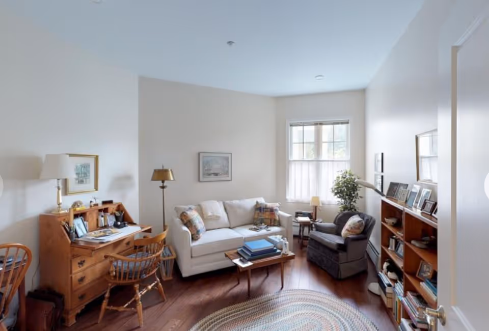 A cozy living room with a white couch, a dark armchair, a wooden coffee table with books on it, a wooden bookshelf filled with books and framed photos, a wooden desk with a chair and a lamp, and a window letting in natural light. The room has light-colored walls and wooden flooring with a round braided rug.