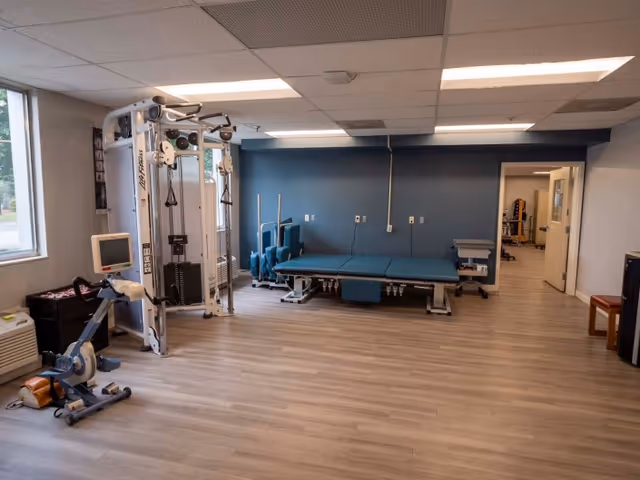 Empty rehabilitation therapy room with exercise equipment, a treatment table, and chairs.