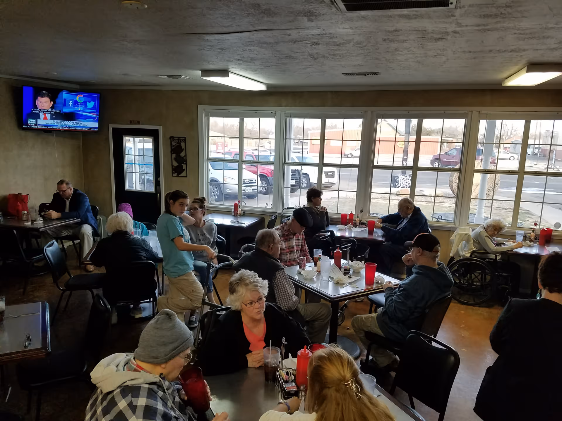 A dining area in an assisted living facility with several elderly residents seated at tables eating and drinking. A young caregiver is assisting one of the residents. Large windows show a parking lot outside, and a television is mounted on the wall.