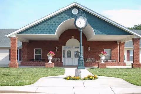 Front exterior view of a brick building with a covered entrance, white double doors, and a decorative clock mounted on a post in front. There are flower pots with pink flowers on either side of the entrance and a well-maintained lawn.