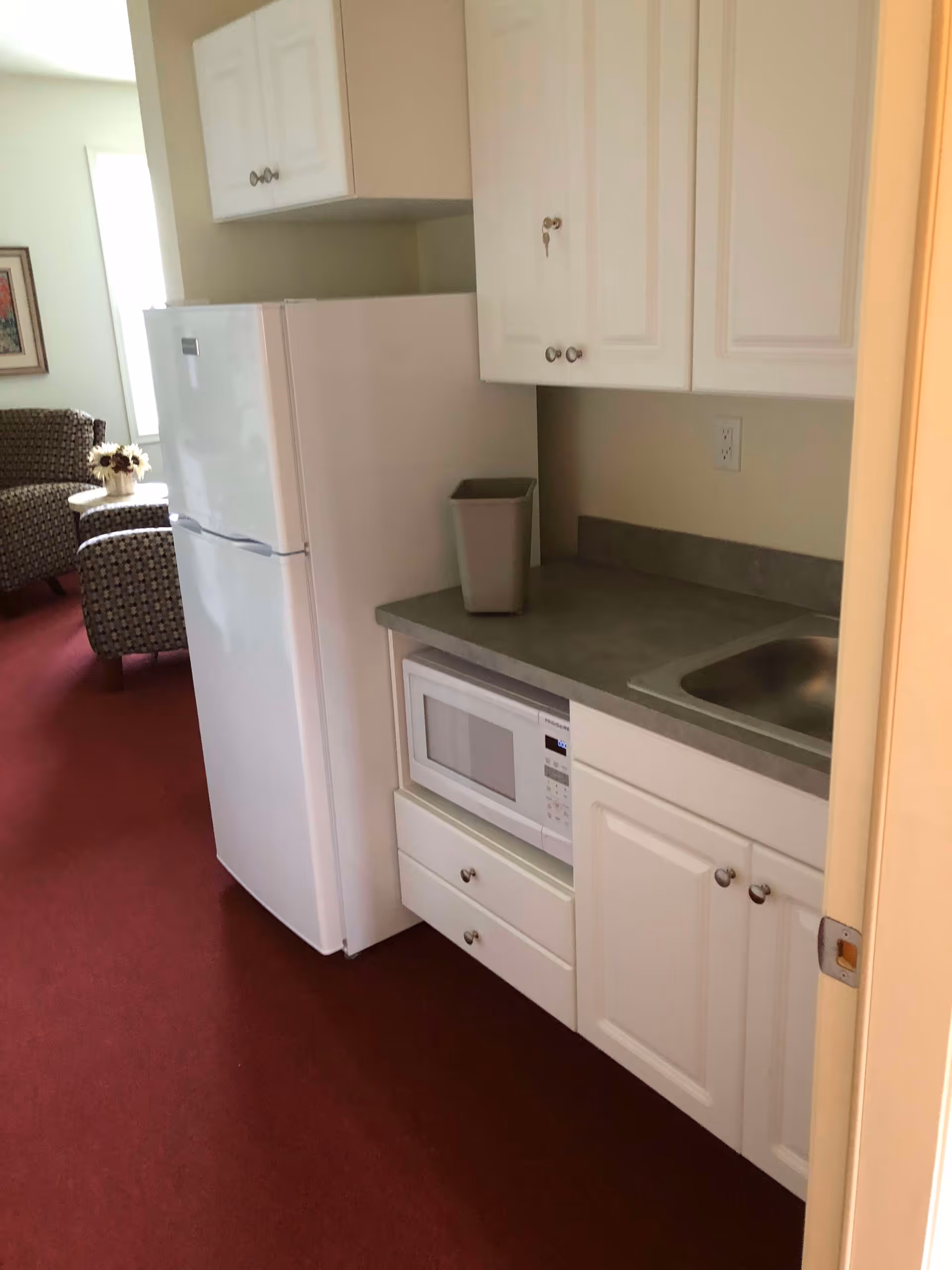 A small kitchenette area with white cabinets, a countertop with a sink, a microwave, and a white refrigerator. In the background, there are two patterned armchairs and a small table with a flower arrangement, all set on a red carpet.