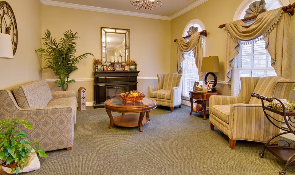 A cozy living room with a patterned sofa on the left, two striped armchairs on the right, a round wooden coffee table in the center with a decorative basket, and a black fireplace with a large mirror above it. The room has beige walls, carpeted floor, two tall windows with draped curtains, a side table with a lamp and framed photos, and several potted plants.