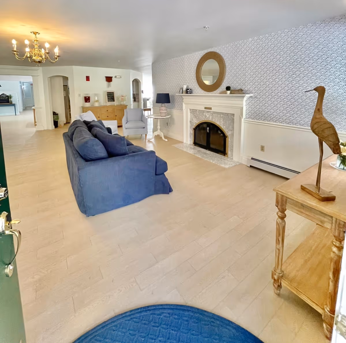 A bright and spacious living room with light wood flooring, a blue sofa, a light blue armchair, a white fireplace with a round mirror above it, a side table with a striped lamp, and a wooden console table with a decorative wooden bird sculpture. The walls have a patterned wallpaper above white wainscoting, and a brass chandelier hangs from the ceiling.