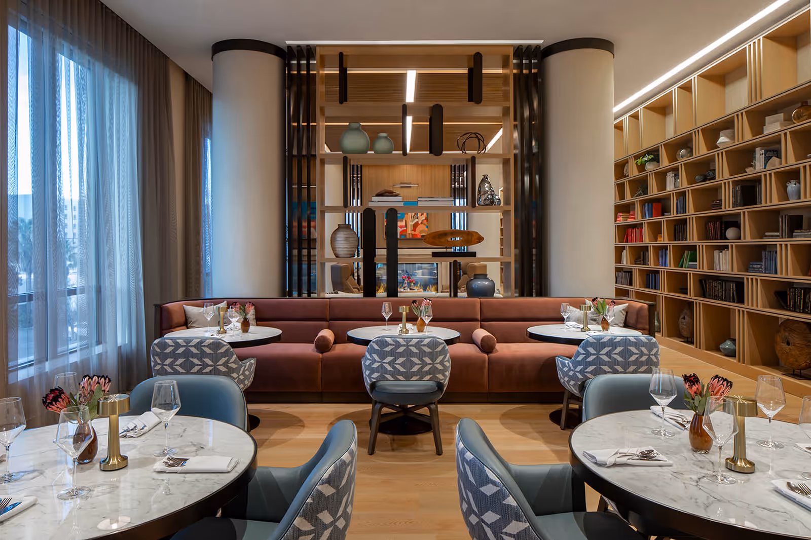 A modern dining area with round marble tables set with wine glasses, napkins, and cutlery. The seating includes patterned blue and gray chairs and a long upholstered bench in a muted pink color. Large windows with sheer curtains allow natural light to fill the room. A wooden shelving unit with decorative vases and books separates the dining area from a cozy lounge space with armchairs and a fireplace. The right wall features a large built-in bookshelf filled with books and decorative items.