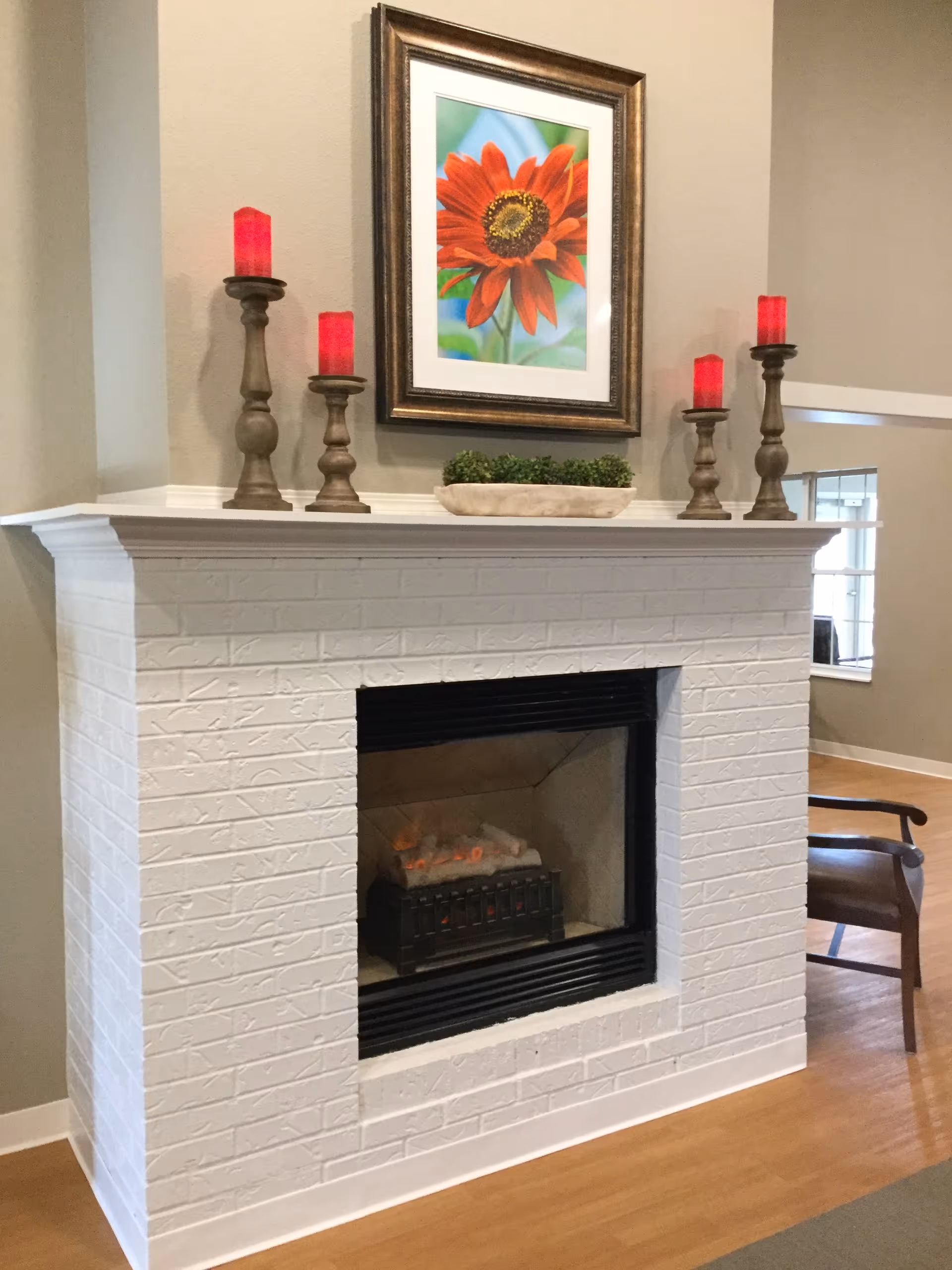 A white brick fireplace with a black firebox containing artificial logs. On the mantel above the fireplace are five candle holders with red candles and a rectangular planter with green plants. Above the mantel hangs a framed picture of a red flower with a green and blue background. To the right of the fireplace is a wooden chair with dark armrests, and a window with white trim is visible in the background.