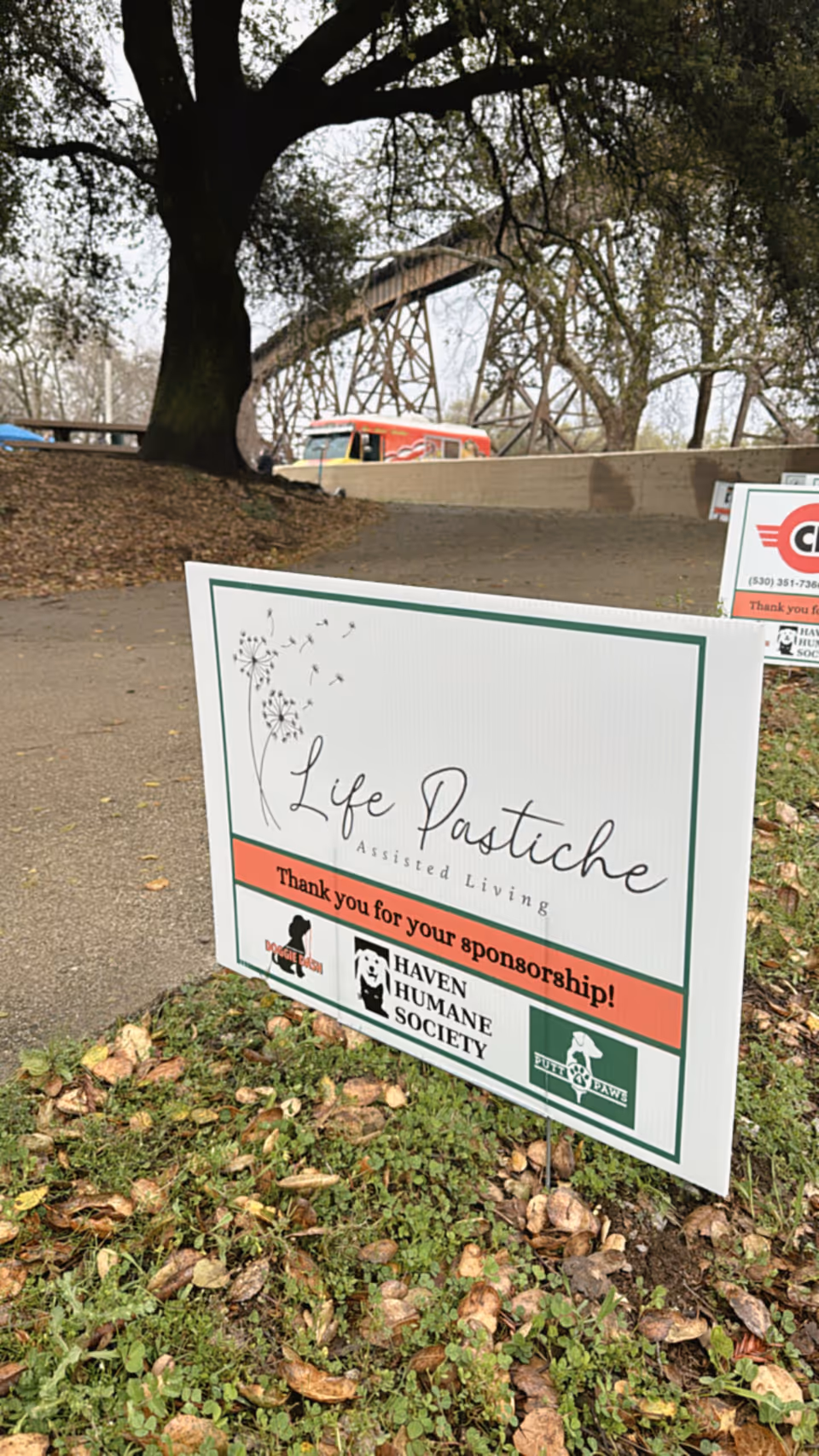 Outdoor scene with a large tree and a pathway. In the foreground, there is a sign on the grass that reads 'Life Pastiche Assisted Living Thank you for your sponsorship!' along with logos for Haven Humane Society and other sponsors. In the background, there is a bridge structure and a red vehicle partially visible.