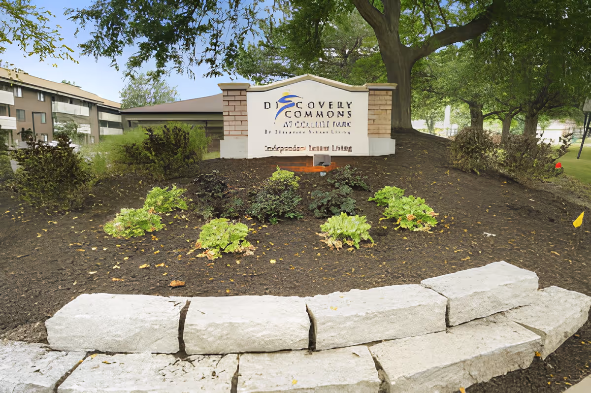 Outdoor view of a landscaped area with a stone border and a sign that reads 'Discovery Commons at College Park' surrounded by trees and bushes, with a multi-story building in the background.