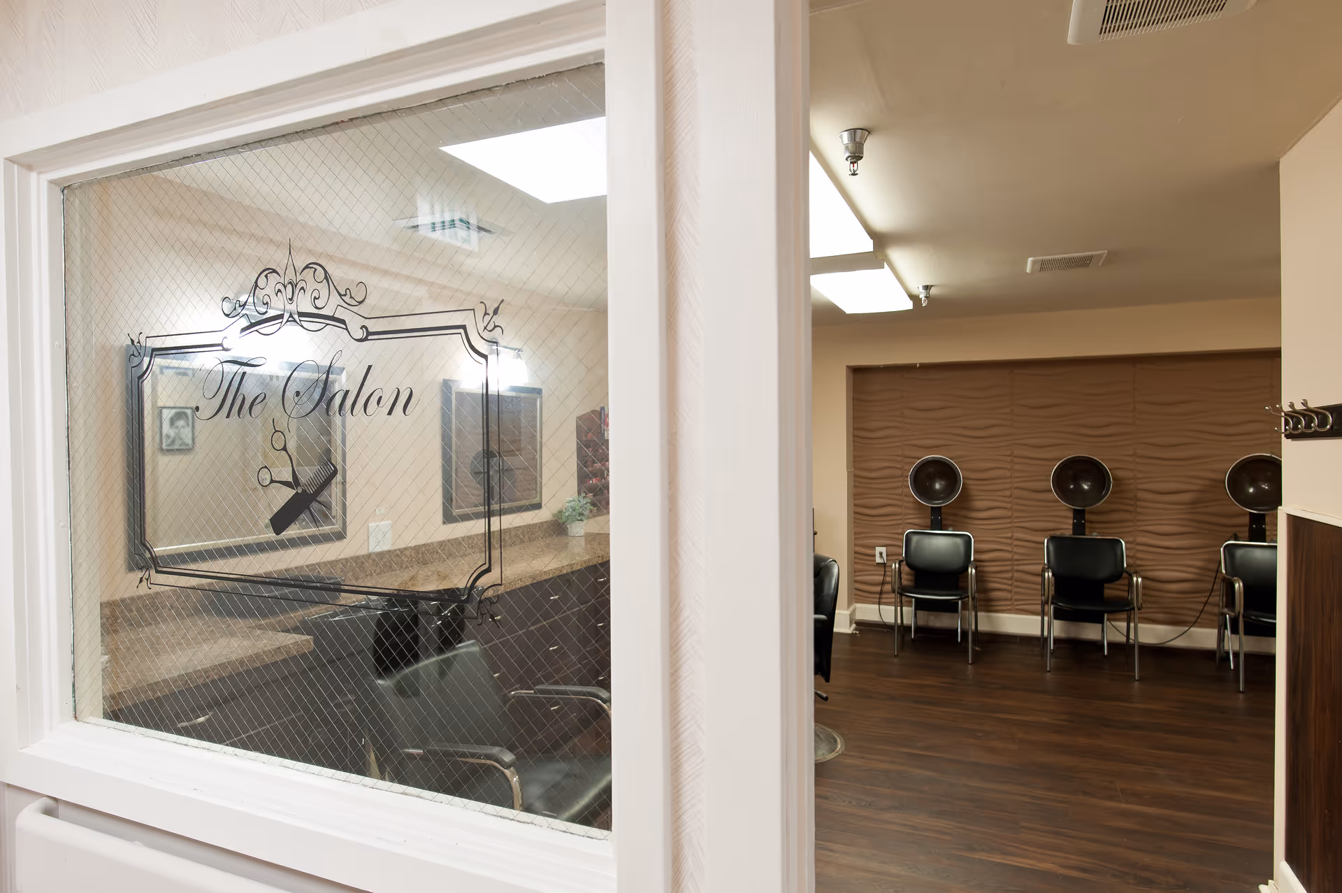 Interior view of a salon room in a senior living facility, showing several black salon chairs with hair dryers against a textured brown wall. The image is taken through a glass window on a door labeled 'The Salon' with decorative scissors and comb graphics.