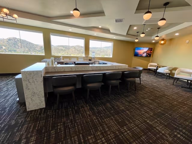 A spacious interior room with large windows showing a view of hills outside. The room features a long marble countertop with several black chairs lined up along it. There are modern pendant lights hanging from the ceiling and a wall-mounted TV on the far wall. Several white cushioned chairs are arranged along the walls.