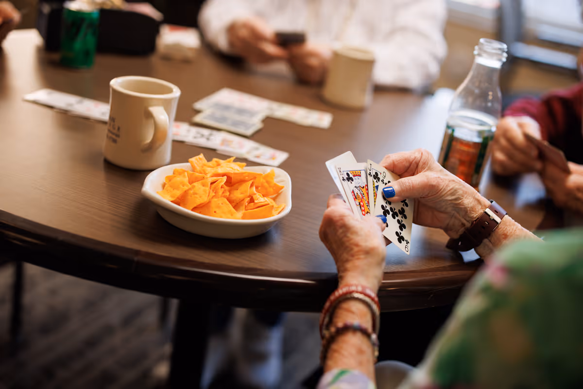 A close-up of elderly hands holding playing cards at a round wooden table with a bowl of tortilla chips, coffee mugs, and a bottle of soda, with other people also holding cards in the background.