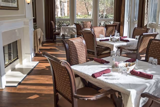 A dining room with several tables covered with white tablecloths and set with glasses, napkins, and a small centerpiece. The room has wooden chairs with patterned upholstery and large windows letting in natural light. There is a fireplace on the left side of the room.
