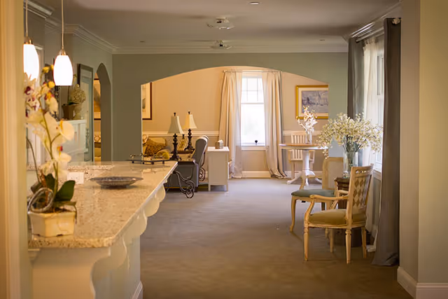 Bright, softly lit common area with a countertop on the left, seating and lamps in the center, and a small dining table by a window with decorative flowers.