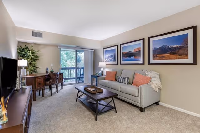 Bright furnished living room with a sofa, coffee table, desk, TV stand and sliding glass door to a balcony.