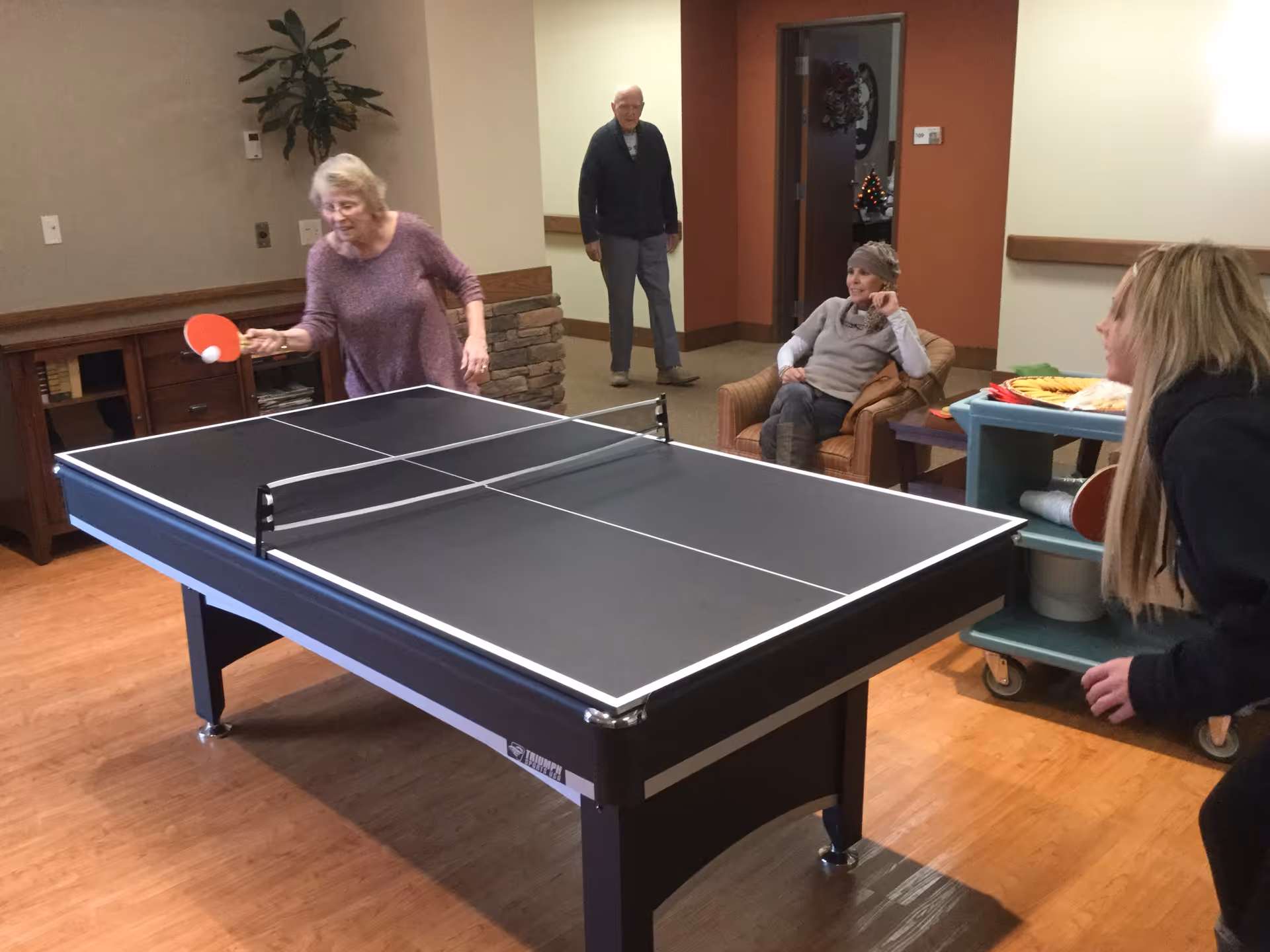 Two women playing ping pong on a table in a common room while a man stands in the background and another woman sits in a chair watching. There is a cart with snacks and a paddle near the seated woman. The room has wooden flooring and neutral-colored walls.