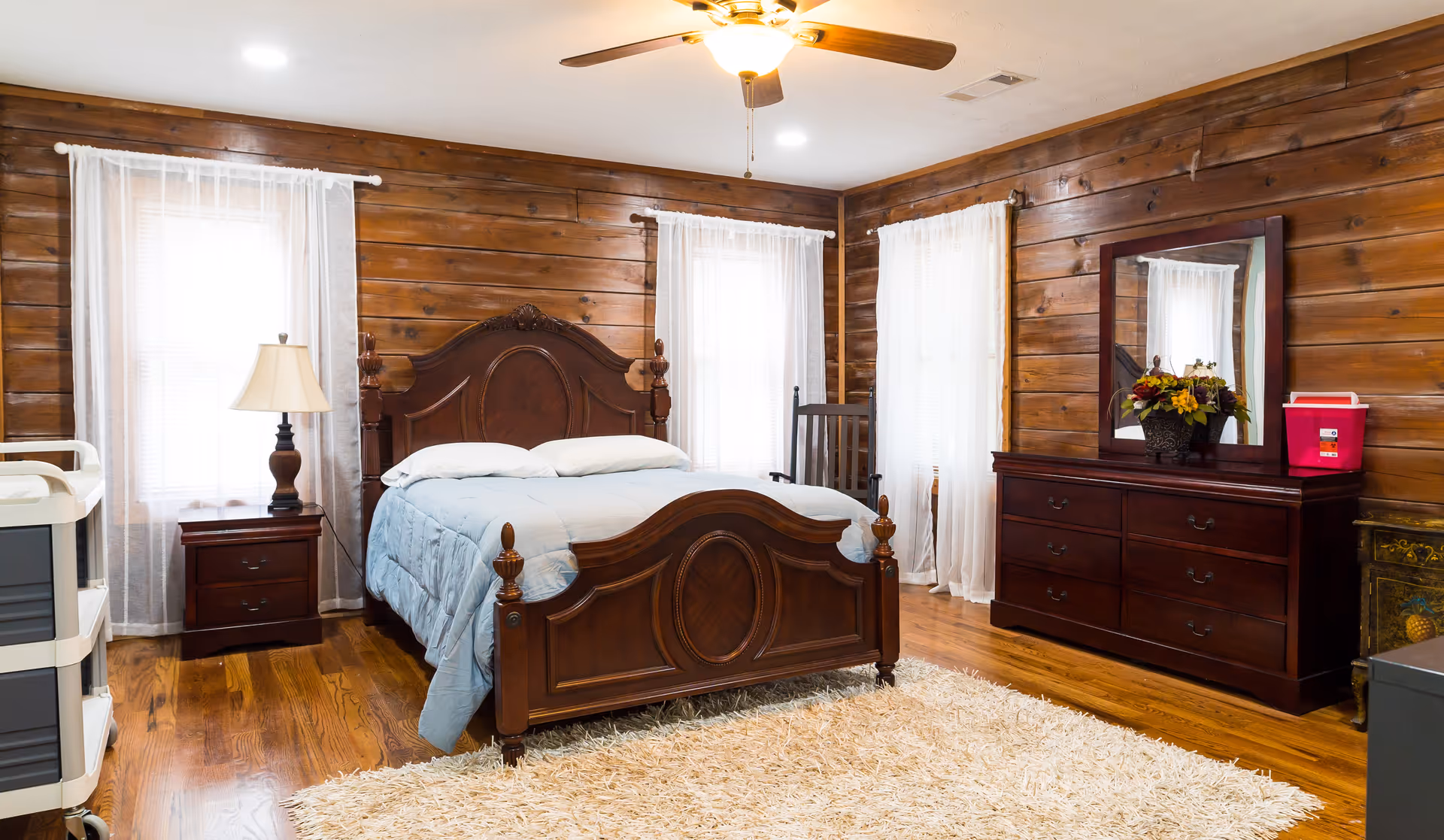A cozy bedroom with wooden paneled walls and hardwood floors. The room features a large wooden bed with a blue comforter and two pillows, a wooden nightstand with a lamp, a wooden dresser with a mirror and a flower arrangement, a rocking chair, and a white shag rug on the floor. Sheer white curtains cover the windows, and a ceiling fan with a light is mounted on the ceiling.